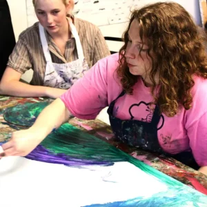 A woman with red hair wearing a pink shirt and an art smock working on a painting