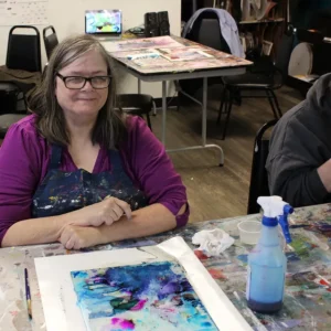 A woman with dark hair and glasses working on a watercolor painting